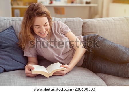Happy Teen Girl Resting on Sofa at the Living Room While Reading her Favorite Novel Book