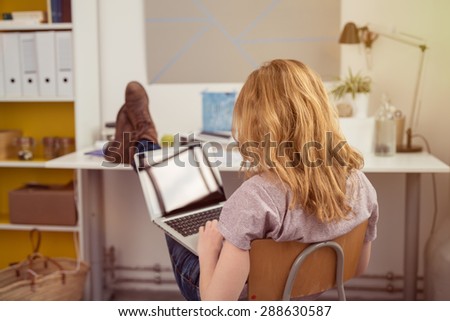 Casual young redhead woman working on a laptop with her feet up on the desk and computer on her lap, view from behind in an office area