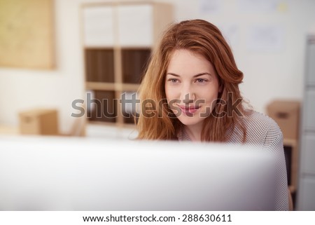 Attractive young woman working on a desktop computer smiling as she leans forwards reading text on the screen, view over the monitor