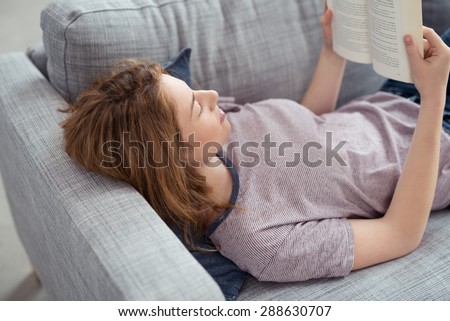 Young woman unwinding reading a book lying on her back on a sofa in the living room as she spends a relaxing day at home