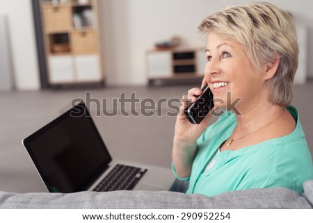 Happy Middle Aged Woman with Laptop Computer, Sitting at the Couch While Talking to Someone on Phone.