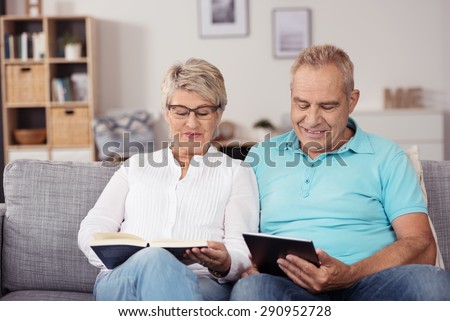 Sweet Middle Aged Couple Reading Something From Book and Tablet Computer While Relaxing at the Living Room Couch