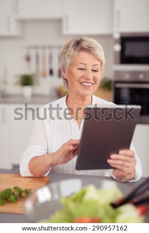 Happy Middle Aged Blond Woman Using her Tablet Computer While Preparing Something to Eat at the Kitchen