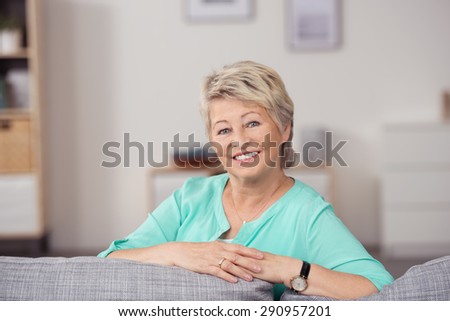 Portrait of a Pretty Senior Blond Woman Sitting on Couch with Arms Leaning on the Pillows, Smiling at Camera