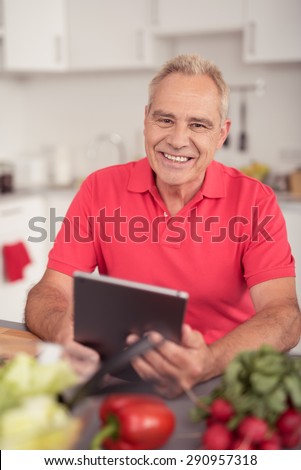 Portrait of a Cheerful Senior Guy Holding his Portable Tablet Computer While at the Kitchen and Looking at the Camera.