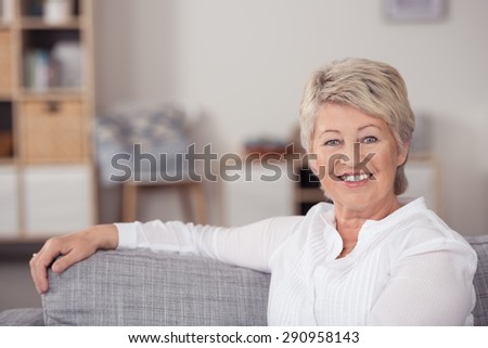 Close up Happy Middle Aged Woman Sitting at the Living Room with One Arm on the Upper Edge of the Sofa, and Smiling at the Camera.