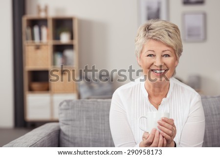 Portrait of a Happy Middle Aged Blond Woman Holding a White Cup While Sitting on the Gray Couch and Smiling at the Camera.
