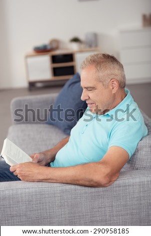 Happy Middle Aged Man Sitting on the Sofa at the Living Room While Reading a Novel Book