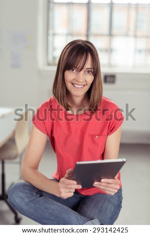 Portrait of a Cheerful Woman in Casual Clothing, Sitting on a Stool While Holding a Tablet Computer and Looking at the Camera.