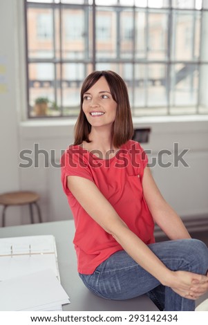 Cheerful Adult Woman in Casual Clothing, Sitting on the Office Table While Holding her Knee Up and Looking Into Distance.