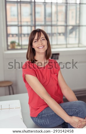 Cheerful Office Woman in Trendy Wear Sitting on Top of a Table with Arms Around her Drawn Up Knee, Looking at the Camera.