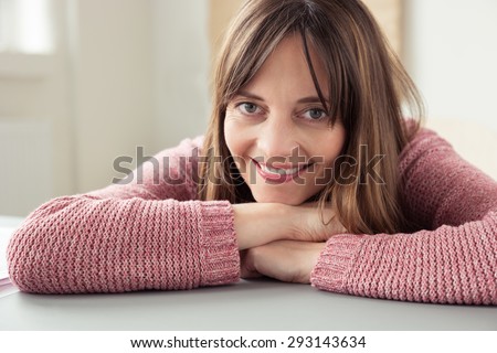 Smiling friendly woman relaxing on a table resting her chin on her folded arms as she looks at the camera, close up of her face