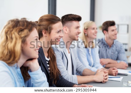 Team of Five Smiling Young Office Workers Sitting at the Boardroom Table, Looking to the Right of the Frame While Listening to Someone.