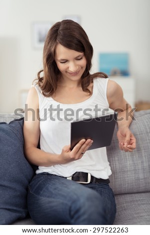 Pretty Young Woman in Casual Outfit Sitting on the Couch, Watching Something on her Tablet Computer with Happy Facial Expression.
