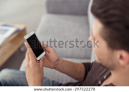 Young Guy Sitting on the Gray Couch at the Living Room, Watching Something on his Mobile Phone