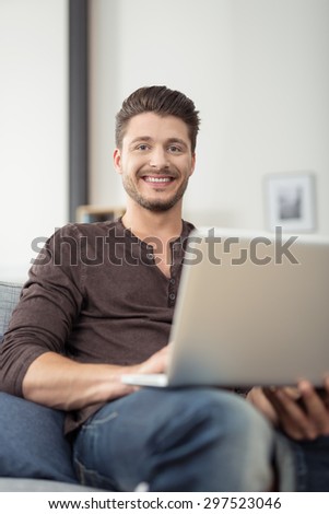 Close up Cheerful Handsome Young Guy Holding his Laptop Computer, Sitting on the Couch at the Living Room and Looking at the Camera.