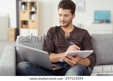 Handsome Young Man Sitting on the Living Room Couch, Writing Some Notes While Looking at his Laptop Computer