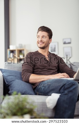 Attractive Young Man Sitting on Couch with Laptop Computer, Looking Into Distance with Happy Facial Expression.