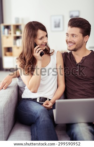 Sweet Young Couple with Laptop Computer Sitting on the Couch, Talking to Someone on Phone with Happy Facial Expressions.