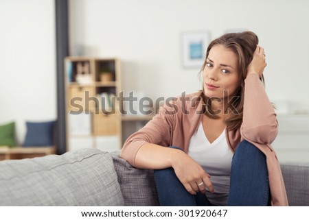 Pretty Young Woman Sitting on Couch In the Living Room with Knees Up and Hand on Head, Looking at the Camera.