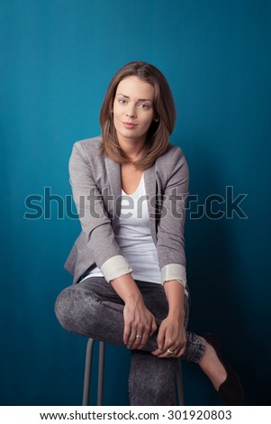 Pretty Young Businesswoman Sitting on a Stool with Legs Crossed and Looking Straight at the Camera Against Blue Green Wall.