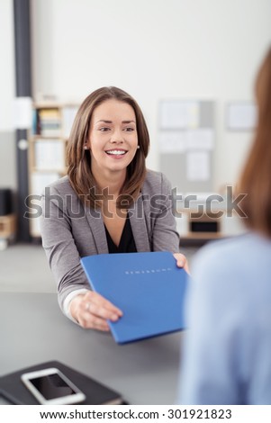 Pretty Young Business Agent Sitting at her Desk Inside the Office, Giving a Blue Folder of Documents to a Client.