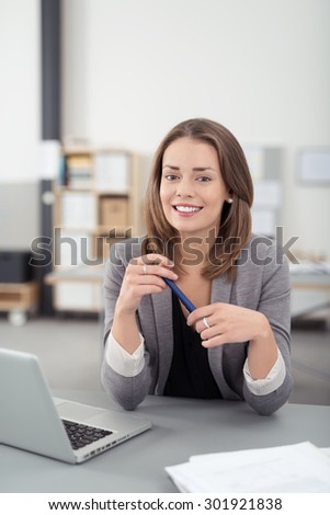 Young Businesswoman Sitting at her Desk with Laptop Computer, Smiling at the Camera While Holding a Pen.