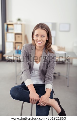 Pretty Young Businesswoman Smiling at the Camera While Sitting on a Stool with Legs Crossed Inside the Office.