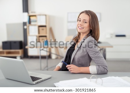 Cheerful Young Office Woman Sitting at her Desk with Laptop Computer, Smiling at the Camera.