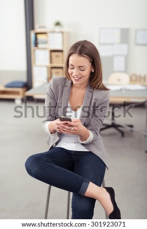 Portrait of a Happy Young Office Woman Sitting on a Stool with Legs Crossed, Busy Texting Someone Using her Mobile Phone