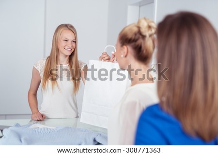 Smiling Young Female Cashier Giving a Shopping Bag to a Female Customer at the Counter Inside a Retail Fashion Store.