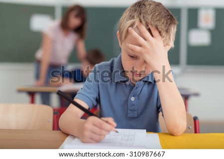 Young schoolboy hard at work in the classroom sitting with his head on his hand reading and writing notes on sheets of white paper