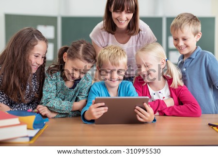 Female teacher with a diverse group of young pupils in class all smiling as they cluster around a tablet computer held by a young boy in the centre