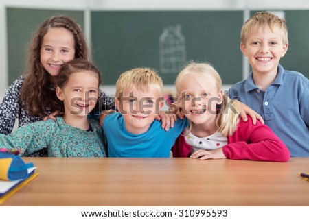 Group of happy grinning pupils in primary school posing arm in arm in a row at a desk looking at the camera, boys and girls