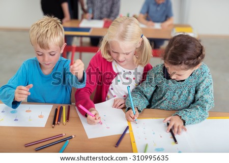 Three young children in kindergarten art class sitting at a desk side by side doodling on sheets of paper with colored pencil crayons, high angle view