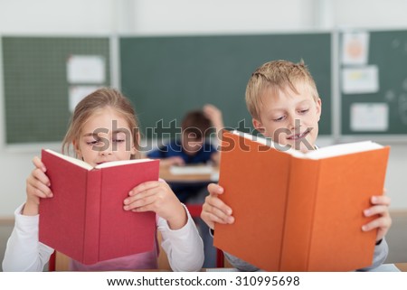 Two young schoolchildren, a boy and girl, sitting side by side at a desk in the classroom reading from textbooks