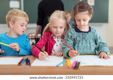 Three young children in kindergarten sharing a desk as they draw with colored pencils on sheets of paper, two little girls and a boy