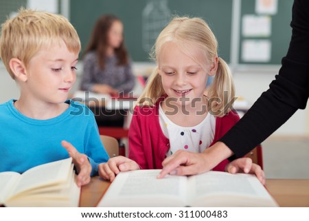 Teacher helping a cute young blond girl at primary school pointing out something in a text book to her as a small boy watches from the side