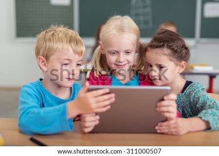 Three young children sharing a tablet computer reading information on the screen as they sit at a desk in the school classroom