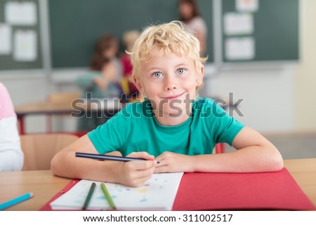 Happy little boy in kindergarten class sitting at his desk drawing with colored pencil crayons and looking at the camera with a beaming smile