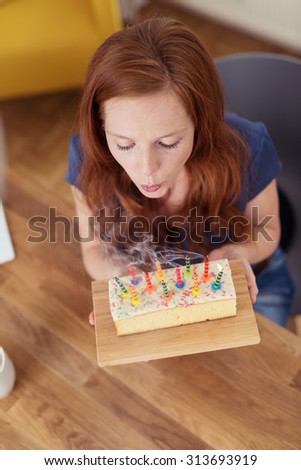 Pretty young redhead woman sitting at a dining table blowing out candles on a cake as she celebrates her birthday, high angle view