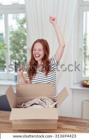 Young woman rejoicing and cheering at the contents of an opened box on the table in front of her raising her hands in the air and laughing, indoors at home