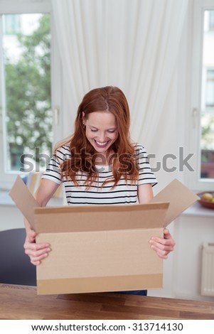 Happy Young Woman Carrying a Carton Box Over a Wooden Table and Looking What is Inside.