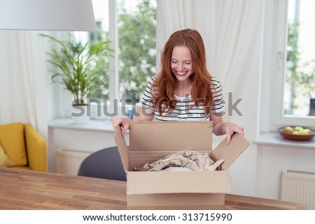 Happy Blond Woman Opening her Present in a Carton box on top of the Wooden Table Inside the House.