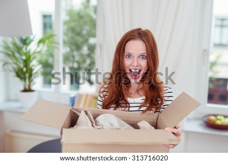 Excited young woman with an opened package in a cardboard box standing with her mouth open in delight and surprise, indoors at home