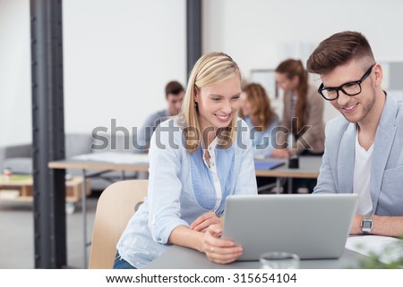 Two Young Office Workers Sitting Inside the Office, Watching Something on Laptop Computer Together with Happy Facial Expression..