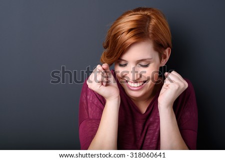 Triumphant happy young woman celebrating raising her fists to her head with a wide ecstatic smile, head and shoulders over a dark studio background