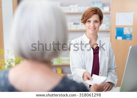 Woman patient handing the pretty young female pharmacist a prescription over the counter, view of the smiling pharmacist over her shoulder