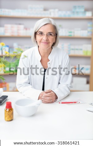 Friendly senior female pharmacist leaning on the counter in the pharmacy smiling at the camera with a bottle of pills and pestle and mortar in front of her
