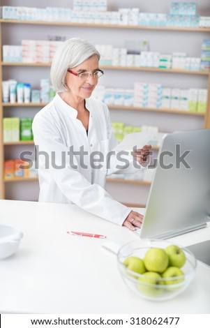 Senior lady pharmacist wearing glasses and a white lab coat working in a pharmacy checking a prescription on the computer